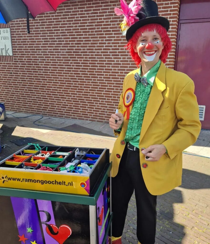 Een vrolijke clown met felrood haar en een gele jas staat naast een kleurrijke kar vol met magische rekwisieten. Zijn geschminkte gezicht straalt met een grote glimlach, en hij draagt een groen shirt, een opvallende staking en een rode hoed versierd met veren. Dit type entertainment is geliefd bij vele goochelshows in Nederland vanwege de speelse interactie en visuele aantrekkingskracht, perfect voor elke feestelijke gelegenheid. Zoek je de leukste goochelaar van Nederland die je evenement onvergetelijk maakt? Dan ben je bij ons aan het juiste adres! Alt tekst: Vrolijke clown naast magische rekwisietenkar tijdens goochelshow in Nederland.