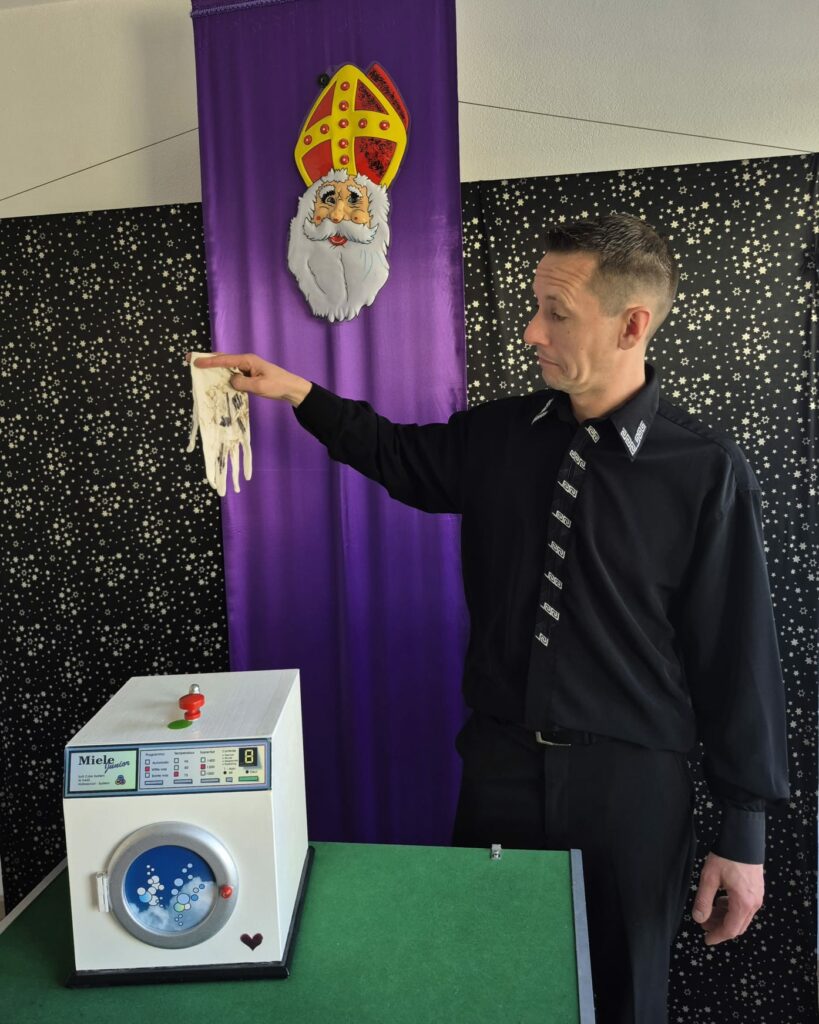 Een vrolijke goochelaar in een zwarte blouse en stropdas houdt een witte goochelhandschoen boven een miniatuur wasmachine, met op de achtergrond een paarse kleur en een grappige afbeelding van Sinterklaas. Deze magische setting straalt precies de speelse sfeer uit van een onvergetelijke Sinterklaasshow voor kinderen. Perfect voor kinderfeestjes, verjaardagen of sinterklaasvieringen met dé leukste kindergoochelaar van Nederland.